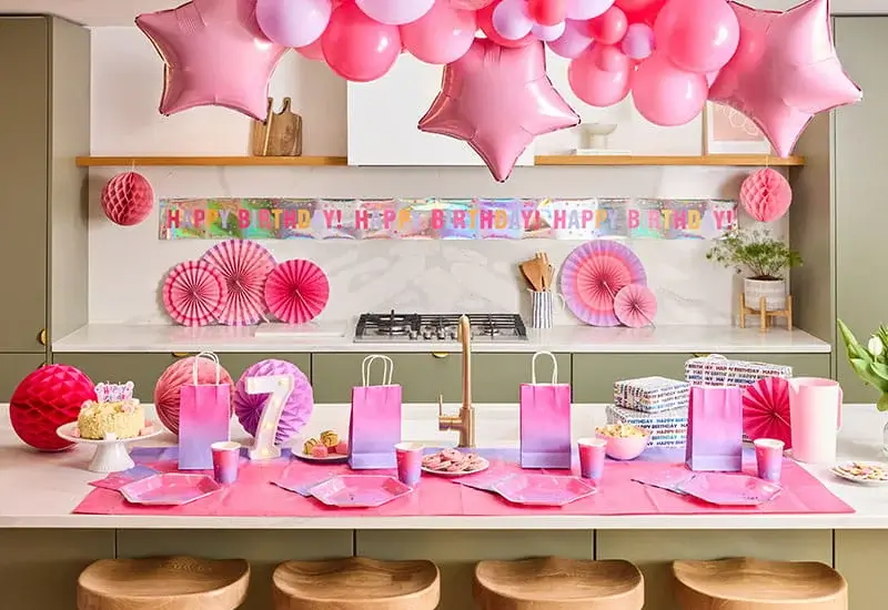 Pink birthday party table with a balloon arch, star balloons, a Happy Birthday banner, gift bags, party tableware, and a lit number 7 decoration.